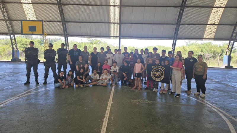 Equipes do Comando de Choque aparecem na fotografia entre os alunos de uma turma do Ensino Fundamental na área do Ginásio de uma Escola. 