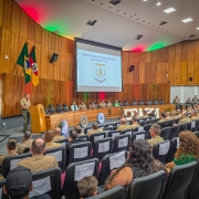O auditório está lotado, o público está sentado assim como os componentes da mesa. Diante do púlpito está o Comandante-Geral da BM em seu discurso. Há luzes com as cores do Estado, vermelha, amarela e verde, iluminando as paredes e o texto do auditório. O brasão do Curso está exposto em um slide do telão.