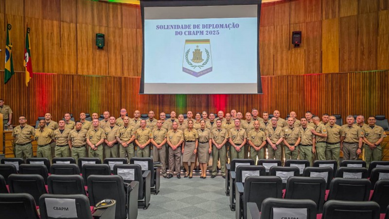 Os PMs formados ocupam o palco dispostos em fileiras. Acima deles, na parede do auditório, está o brasão do Curso exibido no telão. 