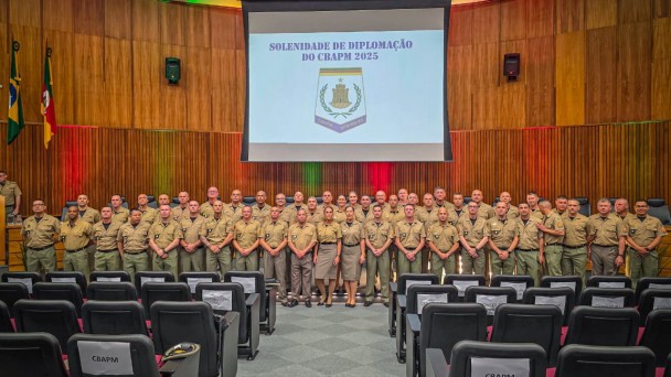 Os PMs formados ocupam o palco dispostos em fileiras. Acima deles, na parede do audit&oacute;rio, est&aacute; o bras&atilde;o do Curso exibido no tel&atilde;o. 