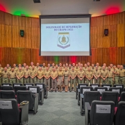 Os PMs formados ocupam o palco dispostos em fileiras. Acima deles, na parede do auditório, está o brasão do Curso exibido no telão. 