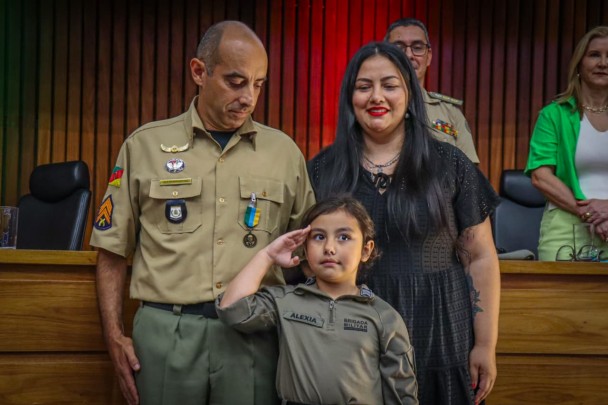 O PM aparece ao lado da esposa e olha para a filha, que está à frente do casal, vestindo uma farda. 