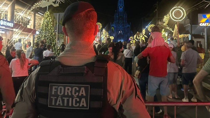 PM está de costas com o colete onde se lê "Força Tática". Ele está de frente ao público que circula em uma rua durante a noite. 