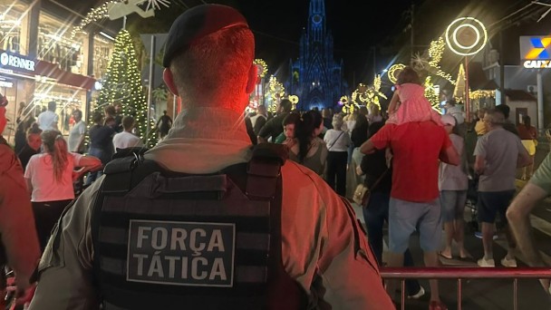 PM est&aacute; de costas com o colete onde se l&ecirc; "For&ccedil;a T&aacute;tica". Ele est&aacute; de frente ao p&uacute;blico que circula em uma rua durante a noite. 