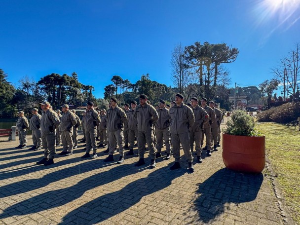 PMs em fileiras aparecem em uma praça da Serra Gaúcha com araucárias ao fundo em um dia ensolarado de inverno. 