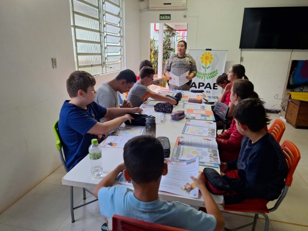 Em sala de aula, a Soldado ministra uma aula do Proerd com adolescentes em volta de uma mesa. Há um caderno de atividades para cada um da turma sobre a mesa, bem como lápis e canetas. 