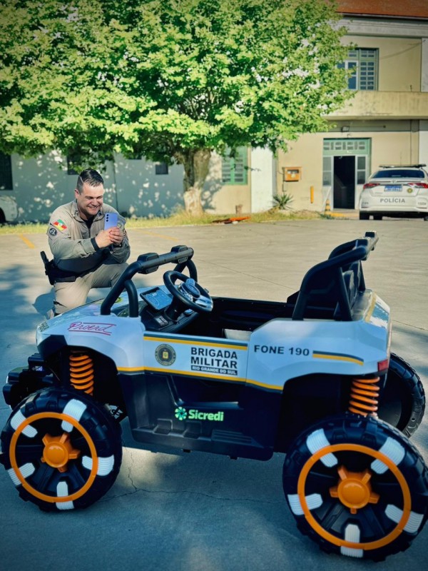 Viatura infantil em destaque com PM a fotografando e uma linda copa de árvore em um dia ensolarado. 