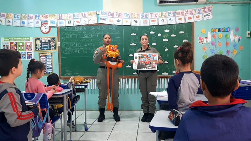 Dupla feminina de PMs diante de uma turma de crian&ccedil;as, segurando um boneco do le&atilde;o s&iacute;mbolo do Proerd e um caderno de atividades. 