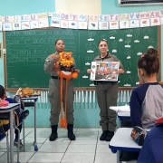 Dupla feminina de PMs diante de uma turma de crian&ccedil;as, segurando um boneco do le&atilde;o s&iacute;mbolo do Proerd e um caderno de atividades. 