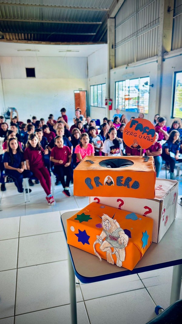 Caixas de perguntas de cor laranja do Proerd facilitam a interação em sala de aula e representam uma das técnicas utilizadas para criar conexão com os estudantes. Ao fundo, aparece a turma. 