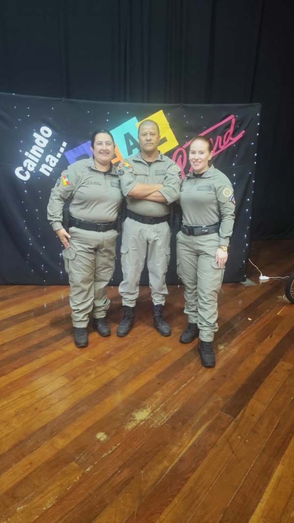 O trio de PMs aparece em fileira em um palco, ao fundo há um banner do Proerd. 