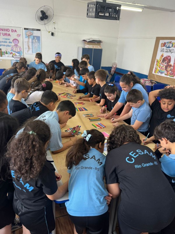 Uma turma está desenhando sobre o papel pardo em cima de uma mesa grande retangular em uma escola. 