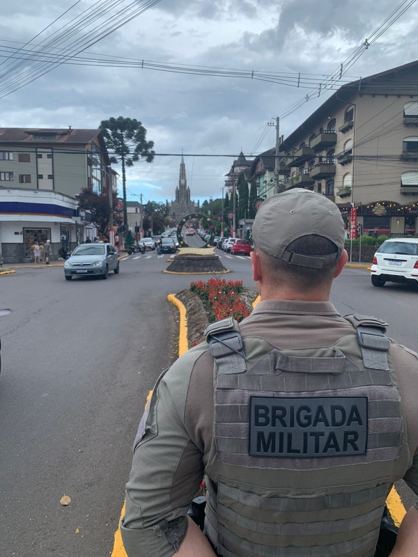 PM masculino eem prontidão na rua diante do canteiro central de uma avenida. Vê-se um veículo de passeio e prédios ao fundo sob o céu nublado. 