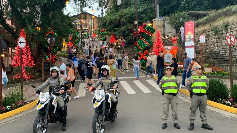 Duas duplas de PMs, uma com motocicletas e outra de prontid&atilde;o, aparecem em um local com grande presen&ccedil;a de p&uacute;blico, durante o dia, decorado com enfeites natalinos. 
