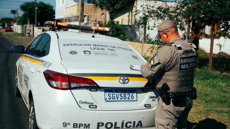 A viatura da BM aparece estacionada em uma rua. Destaca-se o para-brisa traseiro onde se l&ecirc; "Patrulha Maria da Penha Ligue 190". H&aacute; um PM em p&eacute; escrevendo. 