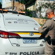 A viatura da BM aparece estacionada em uma rua. Destaca-se o para-brisa traseiro onde se l&ecirc; "Patrulha Maria da Penha Ligue 190". H&aacute; um PM em p&eacute; escrevendo. 