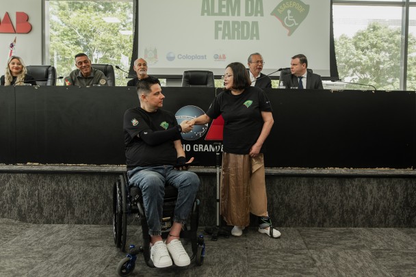 O soldado cadeirante e a presidente em pé têm ao fundo a mesa com os palestrantes do evento "Além da farda"
