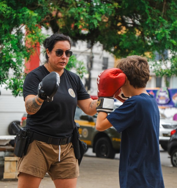 PM feminina aparece ensinando uma técnica de defesa a uma criança em uma área aberta. 