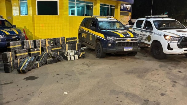 Durante a madrugada, em frente ao posto da PRF, aparecem as viaturas dessa Pol&iacute;cia e da BM. Ao lado delas, est&atilde;o as caixas que representam a grande apreens&atilde;o de maconha. 