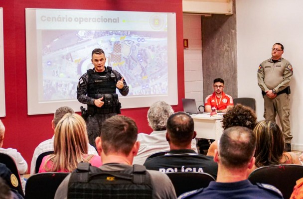PMs falam diante do público presente. Ao fundo a tela que apresenta o cenário operacional da atuação da BM no dia do evento esportivo. 