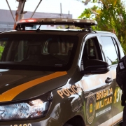 Policial militar do batalh&atilde;o de choque ao lado de uma viatura verde em posi&ccedil;&atilde;o de respeito.