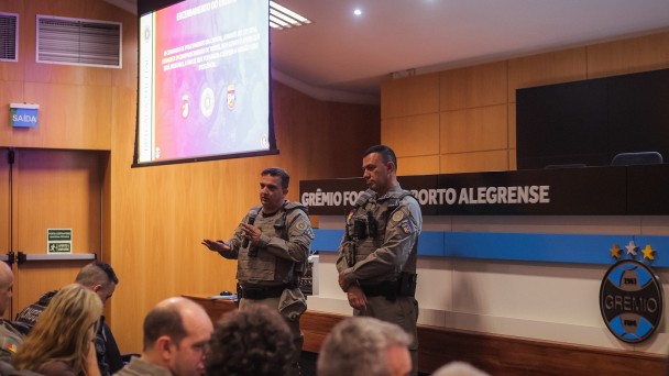 Dupla de comandantes da BM fala diante dos demais representantes de &oacute;rg&atilde;os e institui&ccedil;&otilde;es, com o aux&iacute;lio de um tel&atilde;o, em reuni&atilde;o preparat&oacute;ria.  