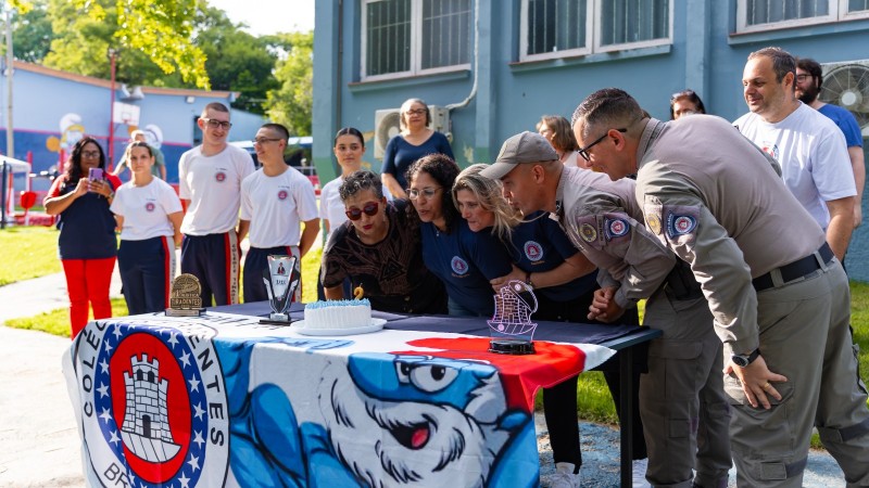 Alunos e professores aparecem pr&oacute;ximo a uma mesa onde tem um bolo de anivers&aacute;rio com uma vela. 