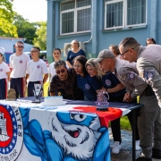 Alunos e professores aparecem pr&oacute;ximo a uma mesa onde tem um bolo de anivers&aacute;rio com uma vela. 