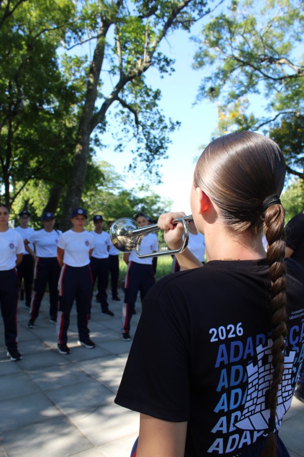 Aluna aparece de costas utilizando um instrumento musical para ensinar as práticas militares aos novos alunos que estão a sua frente.  