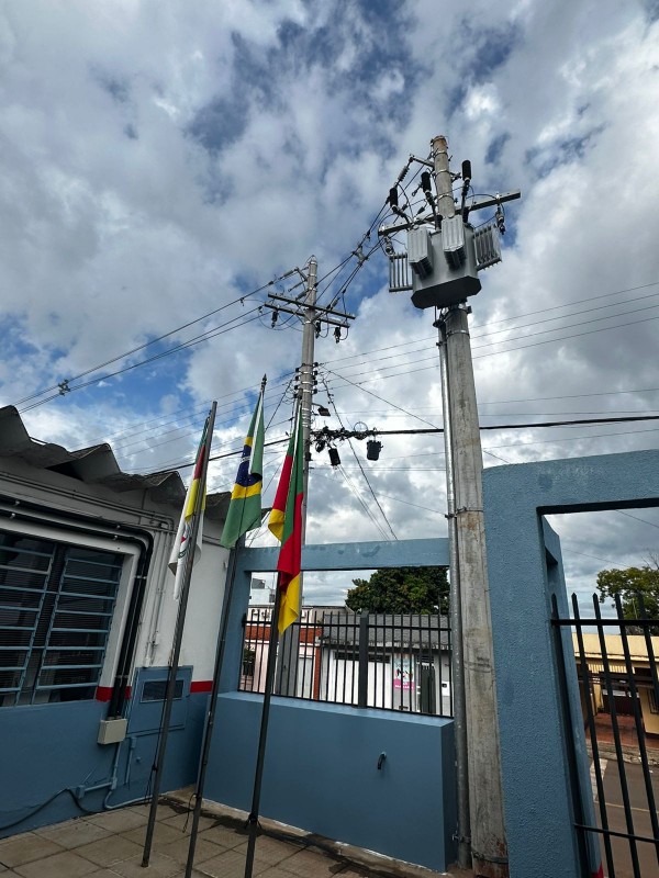 Face interno da entrada do Colégio, destaca-se um poste de energia com um equipamento novo. Ao lado, estão os mastros com as bandeiras. 