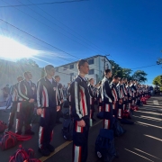 Alunos aparecem em fileiras vestidos com uniformes em ambiente aberto. 