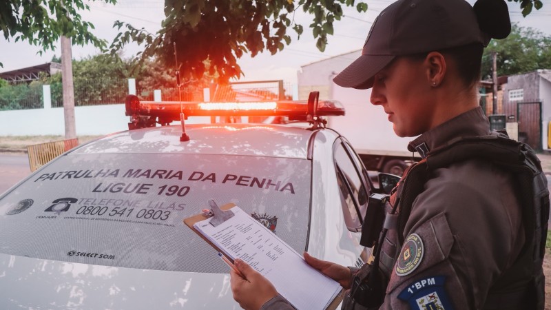 Foto mostra policial militar feminina segurando prancheta em frente a uma viatura da Patrulha Maria da Penha