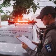 Foto mostra policial militar feminina segurando prancheta em frente a uma viatura da Patrulha Maria da Penha
