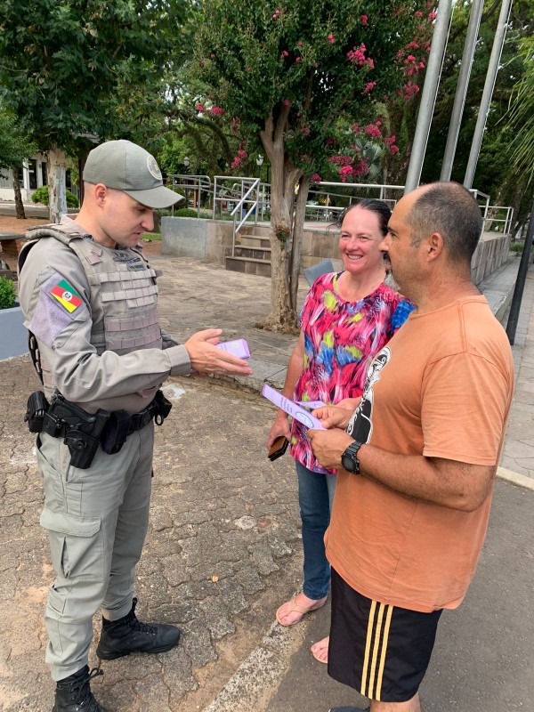 PM conversa com um casal em uma praça municipal e entrega um folder da Patrulha Maria da Penha. 