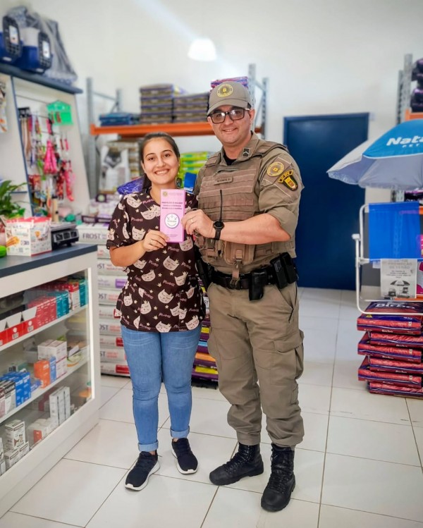 Atendente de uma loja e um PM seguram um folder da Patrulha Maria da Penha da BM. 