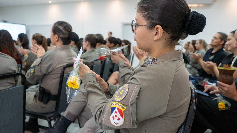 Enquanto as PMs batem palmas, a imagem mostra uma delas, de lado, segurando uma rosa amarela. 