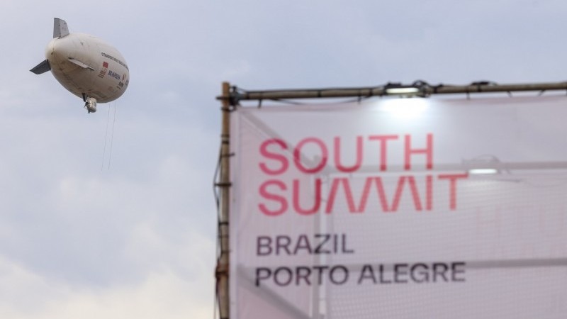 Foto mostra um letreiro com o nome South Summit Brazil Porto Alegre. Ao lado dele, um dirig&iacute;vel.