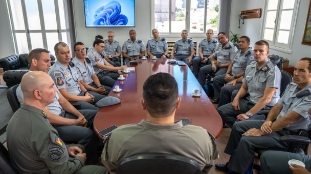 A foto mostra praticamente todos os integrantes da reuni&atilde;o da comitiva com o Comando-Geral da BM. Todos os PMs usam farda e sobre a mesa de trabalho est&atilde;o x&iacute;caras de caf&eacute; e aparelhos celulares. 