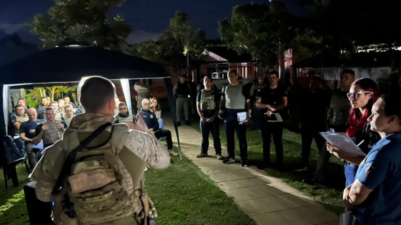 Foto mostra uma tenda com alunos em um jardim. Na frente, e em frente &agrave; c&acirc;mera, est&aacute; um militar fardado do Bope fazendo debriefing