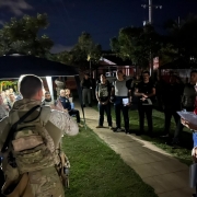 Foto mostra uma tenda com alunos em um jardim. Na frente, e em frente &agrave; c&acirc;mera, est&aacute; um militar fardado do Bope fazendo debriefing