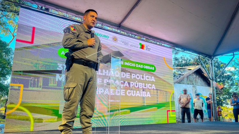 Comandante-Geral fala ao p&uacute;blico com um microfone. Est&aacute; de p&eacute; em um palco e ao fundo h&aacute; um tel&atilde;o com informa&ccedil;&otilde;es do evento. 