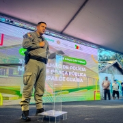 Comandante-Geral fala ao p&uacute;blico com um microfone. Est&aacute; de p&eacute; em um palco e ao fundo h&aacute; um tel&atilde;o com informa&ccedil;&otilde;es do evento. 