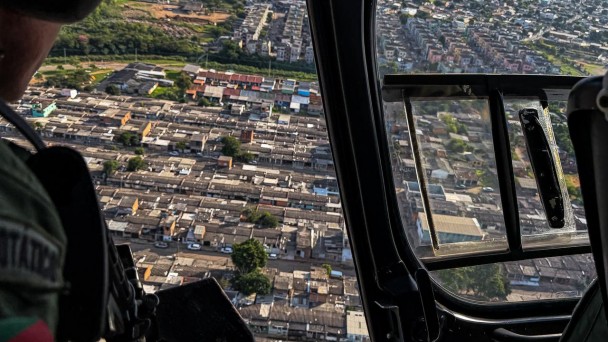 Imagem a&eacute;rea de um bairro feita do interior de um helic&oacute;ptero. Aparece um PM armado no lado esquerdo. 