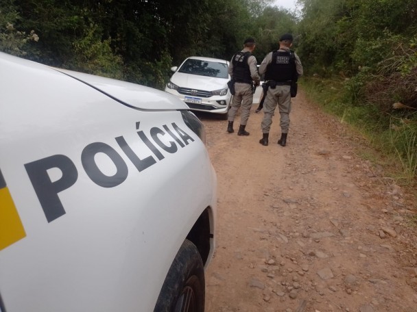 Viatura em primeiro plano e, à frente, dois PMs aparecem em pé na estrada de chão próximos a um veículo branco no canto superior esquerdo. 