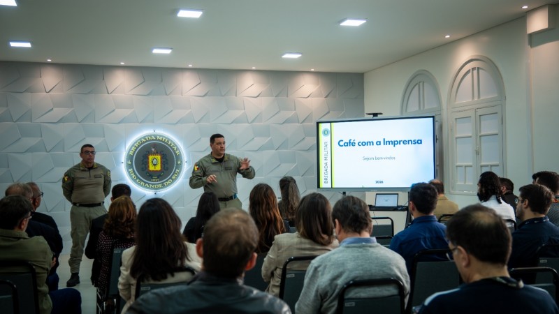 Os Coron&eacute;is est&atilde;o em p&eacute; diante dos jornalistas que ocupam as cadeiras do audit&oacute;rio da sede da BM em Porto Alegre. H&aacute; uma tela de apresenta&ccedil;&atilde;o de slides e o s&iacute;mbolo da Institui&ccedil;&atilde;o ao centro da imagem. 