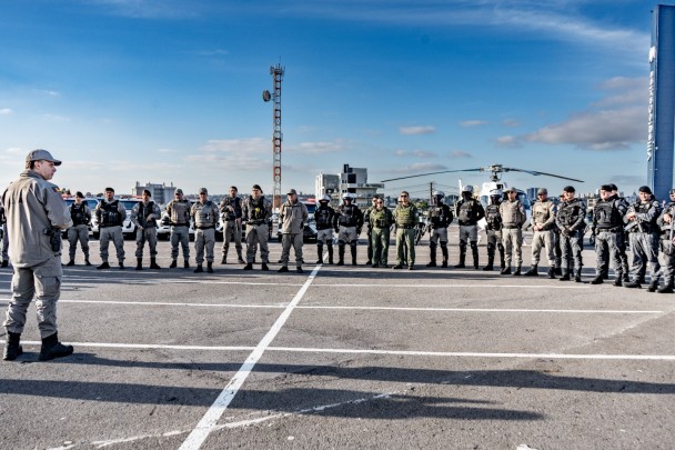Foto mostra policiais militares reunidos ao redor do comandante, que está em frente a eles, dando a preleção.
