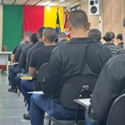 Em um auditório da Brigada Militar, dezenas de candidatos ao Curso Superior de Polícia Militar estão sentados em fileiras, de costas para a câmera, acompanhando orientações. Ao fundo, aparecem as bandeiras do Rio Grande do Sul e do Brasil, além do brasão da Instituição.