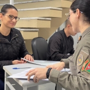 Uma servidora da Brigada Militar realiza atendimento individual a uma candidata durante a conferência de documentos. Ambas estão sentadas frente a frente em uma mesa, em ambiente interno da Corporação.