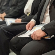 A imagem mostra uma fileira de policiais militares sentados durante uma solenidade. Em destaque, aparecem as m&atilde;os de um dos participantes segurando um certificado, enquanto outros formandos, trajando terno escuro, permanecem alinhados ao fundo. A fotografia utiliza foco seletivo, evidenciando os documentos recebidos pelos concluintes do curso.