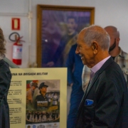 Fotografia colorida mostra o coronel da reserva remunerada Jerônimo Carlos Santos Braga sorrindo durante a visita à exposição. Ao fundo aparecem painéis informativos e outros convidados circulando pelo espaço do Museu da Brigada Militar.